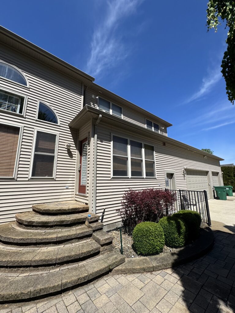 Front exterior of a beige siding house with arched windows and a circular stone staircase leading to the door under a bright blue sky.