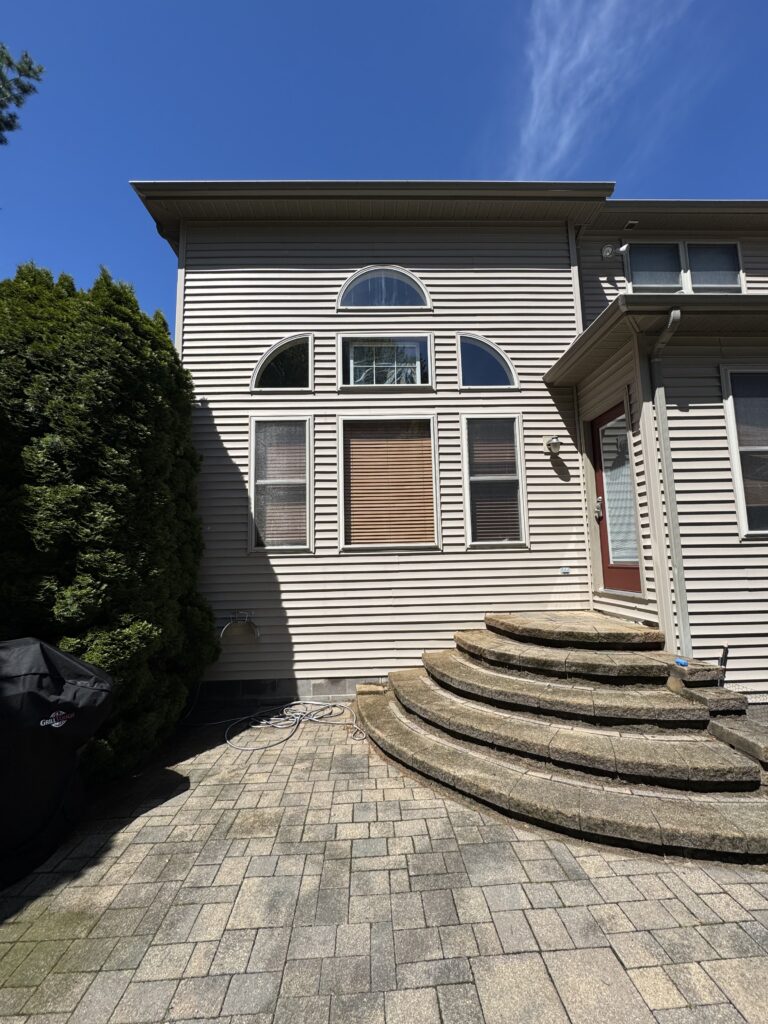 Front view of a beige siding house with arched and rectangular windows, curved stone steps leading to a red door, and a large green hedge on the left under a blue sky.