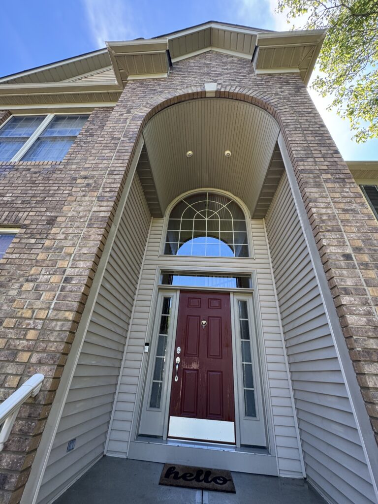Red front door under a brick arched entryway with a transom window, beige siding, and a 'hello' doormat in the doorway.