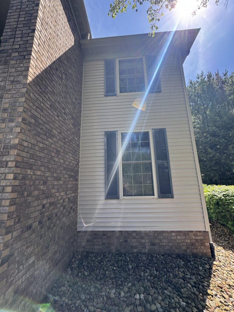 Sunlit corner of a beige siding house with blue shutters and a brick foundation, stacked with gravel around the base.