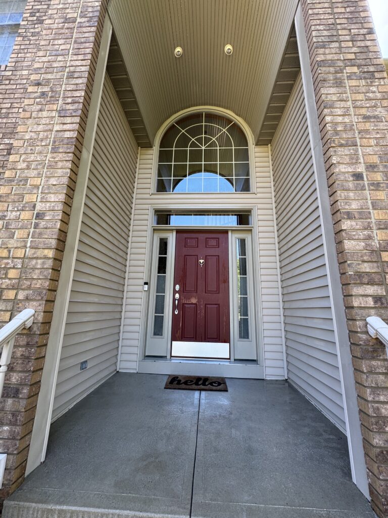 Red front door with glass sidelights and arched transom window, framed by beige siding and brick columns at a home entry.