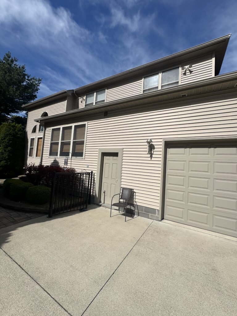 Beige two‑story townhouse with a large front window, garage, and a small chair on the driveway under a blue sky.