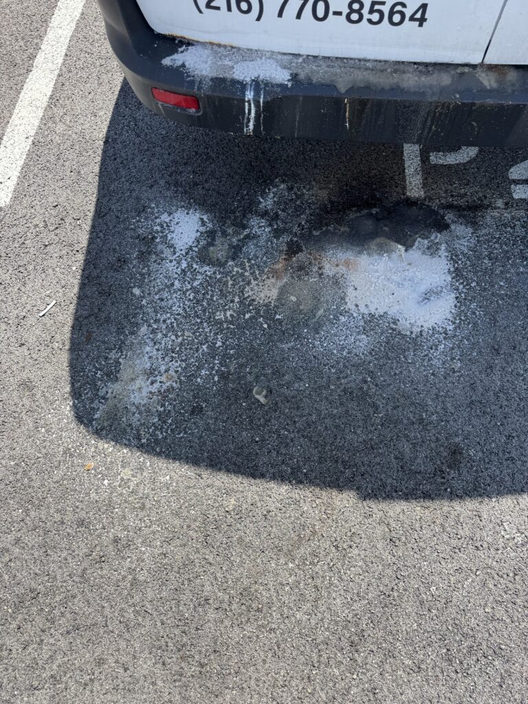 Rear of a parked vehicle with a dirty bumper and white salt/ice residue along the bottom edge, spills and a wet patch on the asphalt beneath it in a parking spot.