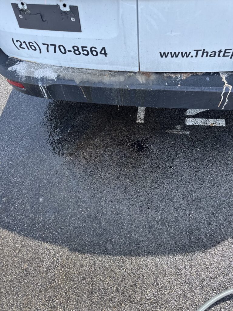 Rear view of a light-blue van showing the contact number (216) 770-8564 and part of a website URL, with a dirty bumper and wet asphalt nearby.