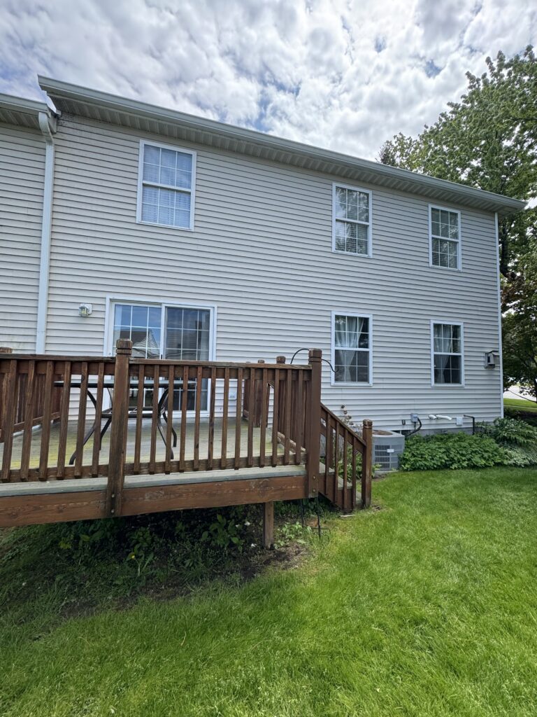 Back of a two-story beige house with white trim, multiple windows, and a wooden deck railing in the yard.