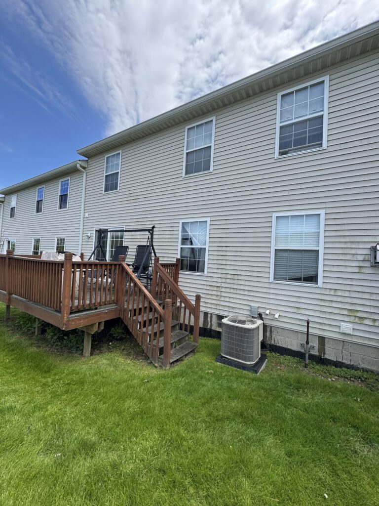 Backyard of a beige-siding house with a wooden deck, outdoor seating, and an air conditioner unit on the ground beside a green lawn under a blue sky with clouds.