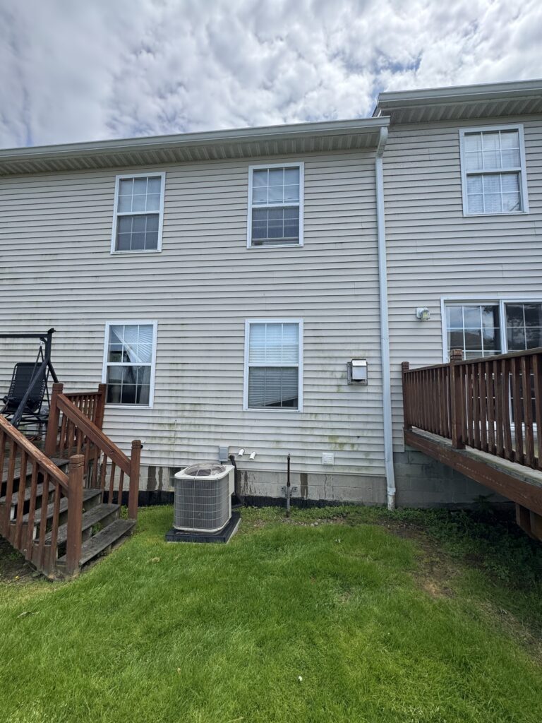 Backyard view of a beige townhouse with vinyl siding, showing multiple windows, a wooden deck, and an outdoor air conditioner unit on the lawn.
