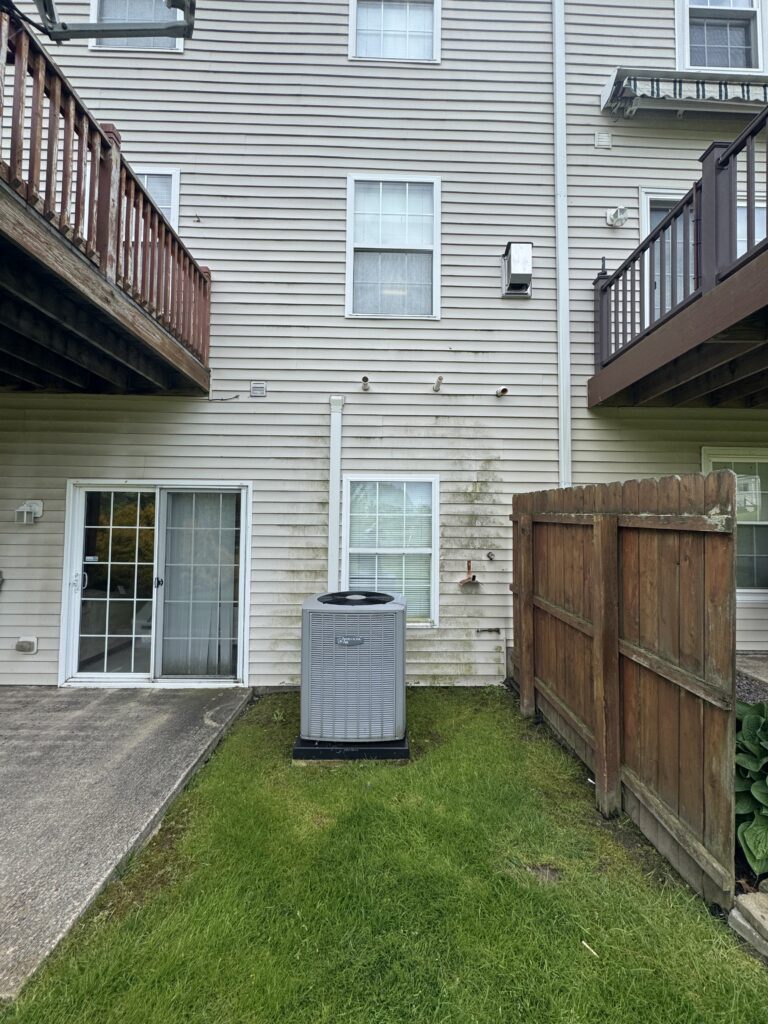 Backyard of a beige townhouse with an outdoor AC unit on a small grassy patch beside a sliding glass door and a wooden fence on the right.