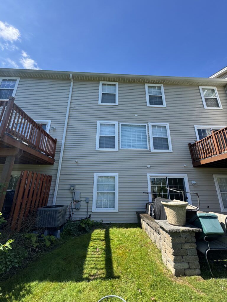 Rear view of a two-story beige siding apartment building with white-framed windows and two wooden decks on either side, a grassy yard, and a stacked stone planter in the foreground under a clear blue sky.