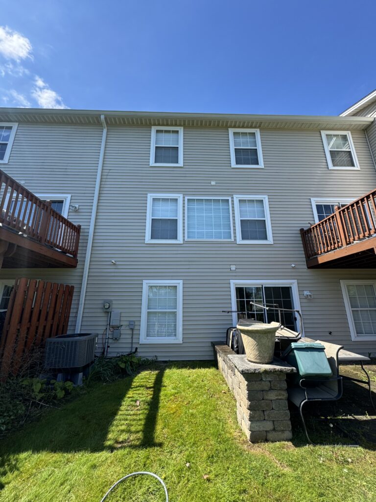 Rear view of a beige, two-story townhouse with multiple white-framed windows and wooden balconies on each side, under a blue sky (informative).