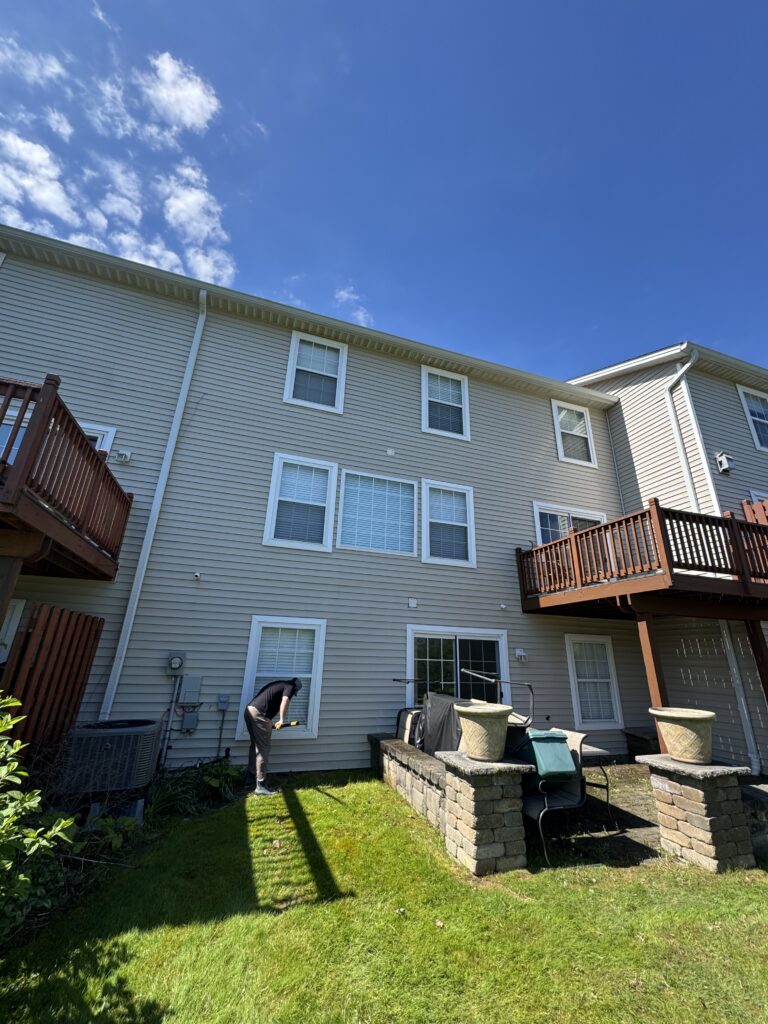 Backyard scene of a beige townhouse with several windows; a person in dark clothing is kneeling by a door and working near the wall, lawn and deck visible outdoors.
