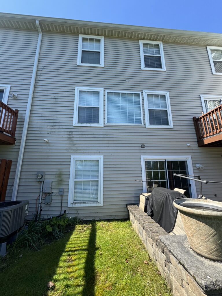 Back of a beige townhouse with white-framed windows, a wooden deck on the right, and a grassy yard with a covered grill nearby.