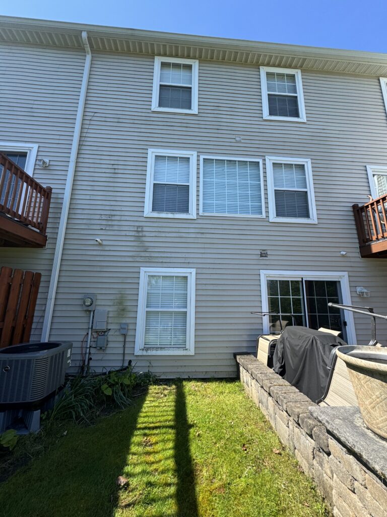 Backyard view of a beige townhouse with white-framed windows, deck rails on both sides, and a low stacked-stone wall.
