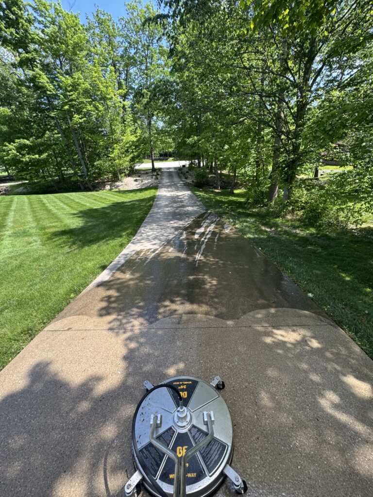 First-person view down a concrete park path lined with green trees, with a large circular lawn tool in the foreground.