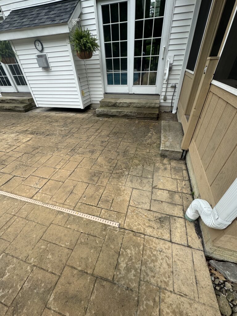 Backyard patio with tan pavers and stone steps leading up to white-framed French doors, adjacent to beige siding and a downspout.