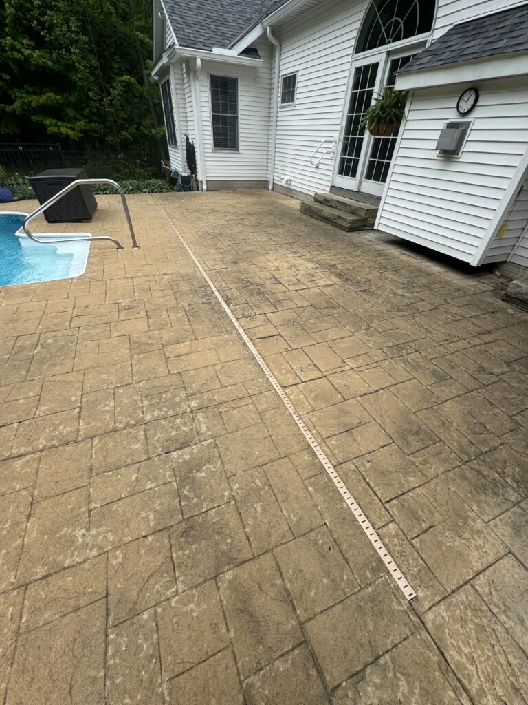 Patio area beside a white house with French doors, a blue pool on the left, and a long measuring tape running across tan brick pavers.