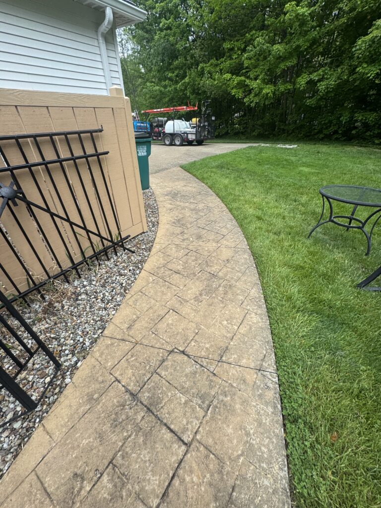 Curved stamped concrete path beside a green lawn, with a beige wooden wall and a leaning black metal railing on the left, and a trailer in the driveway beyond trees.
