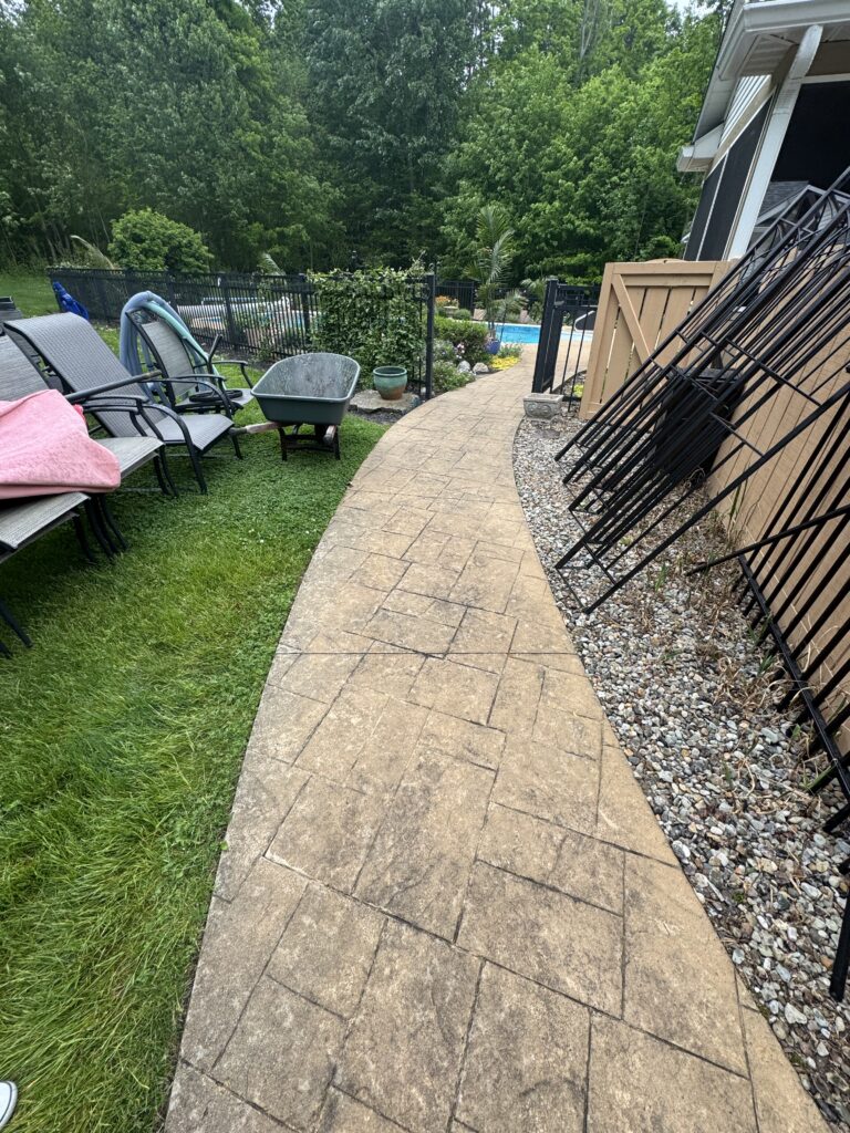 Curved stamped concrete walkway in a backyard, with stacked patio chairs on the left, a gray wheelbarrow, and a view of a pool beyond a black fence.