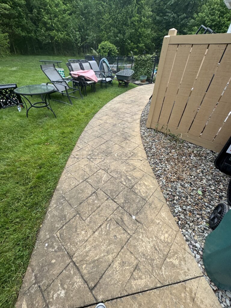 Backyard patio with a curved stamped concrete path, multiple metal chairs, and a glass table on the grassy side.
