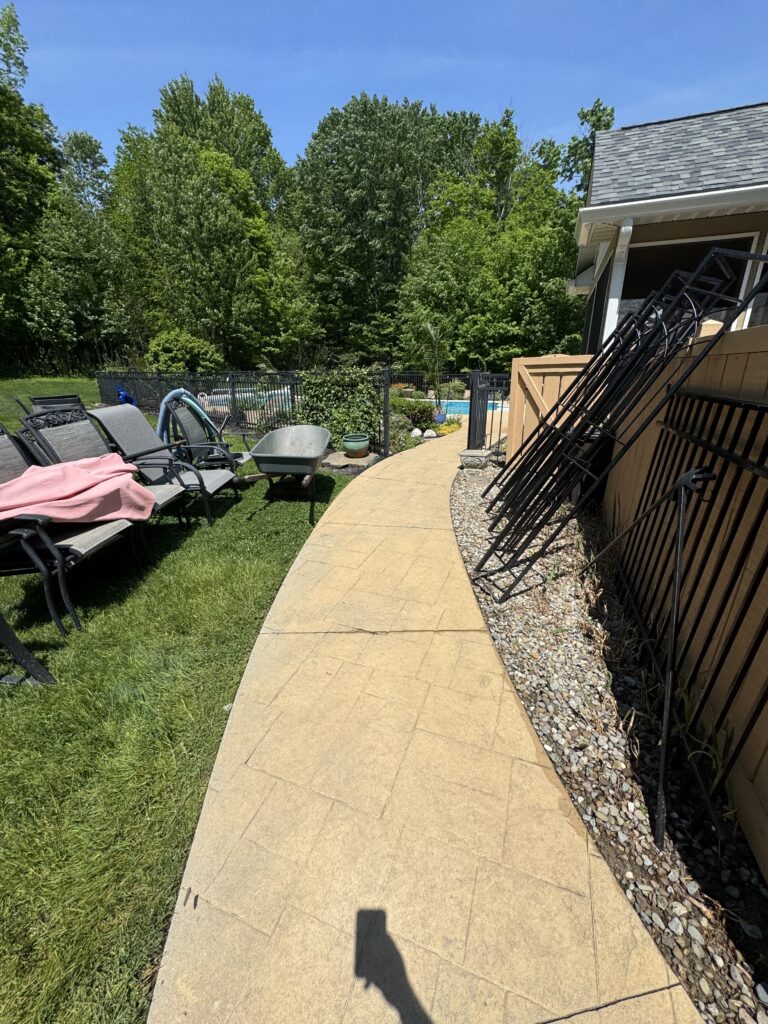 Backyard patio with beige paver path, stacked black metal chair frames along a wooden fence, and a lawn with a pool and trees in the background on a sunny day.