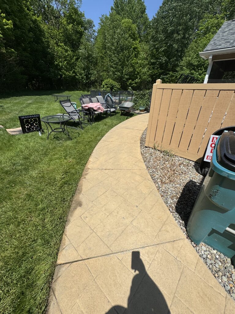 Curved stone patio with outdoor seating including black metal chairs and glass table, facing a grassy yard with trees in the background.