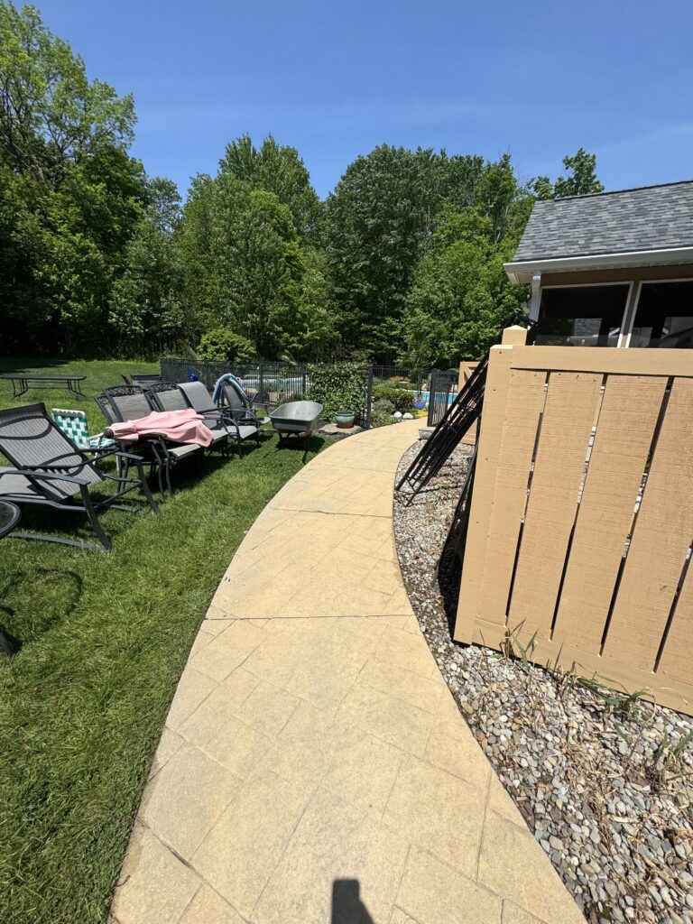 Curved beige stone pathway with gravel border, brown privacy slats on the right, and patio chairs facing a fenced pool in the background.