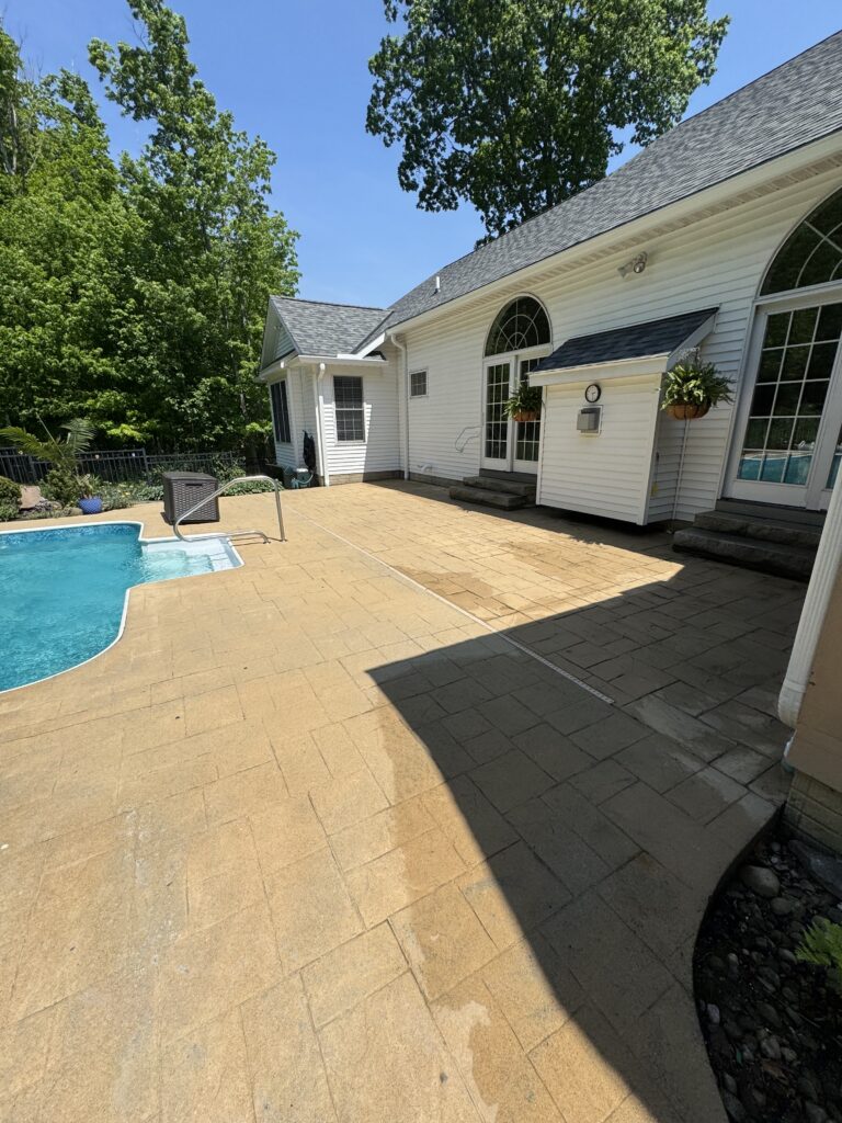 Backyard scene with a white house, arched window, and a rectangular in-ground pool on a sunny day, surrounded by trees and a tan brick patio.