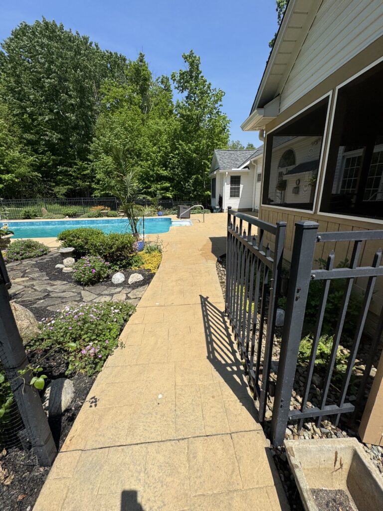 Poolside patio with a blue rectangular pool, stone walkway, and landscaped garden under a bright blue sky.