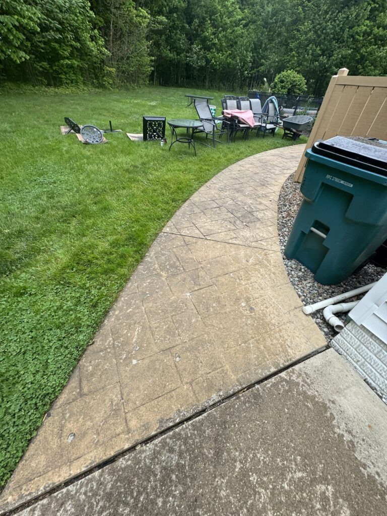 Backyard scene with a curved stone patio and a cluster of outdoor chairs on the grass; a blue recycling bin is at right and miscellaneous debris sits nearby.