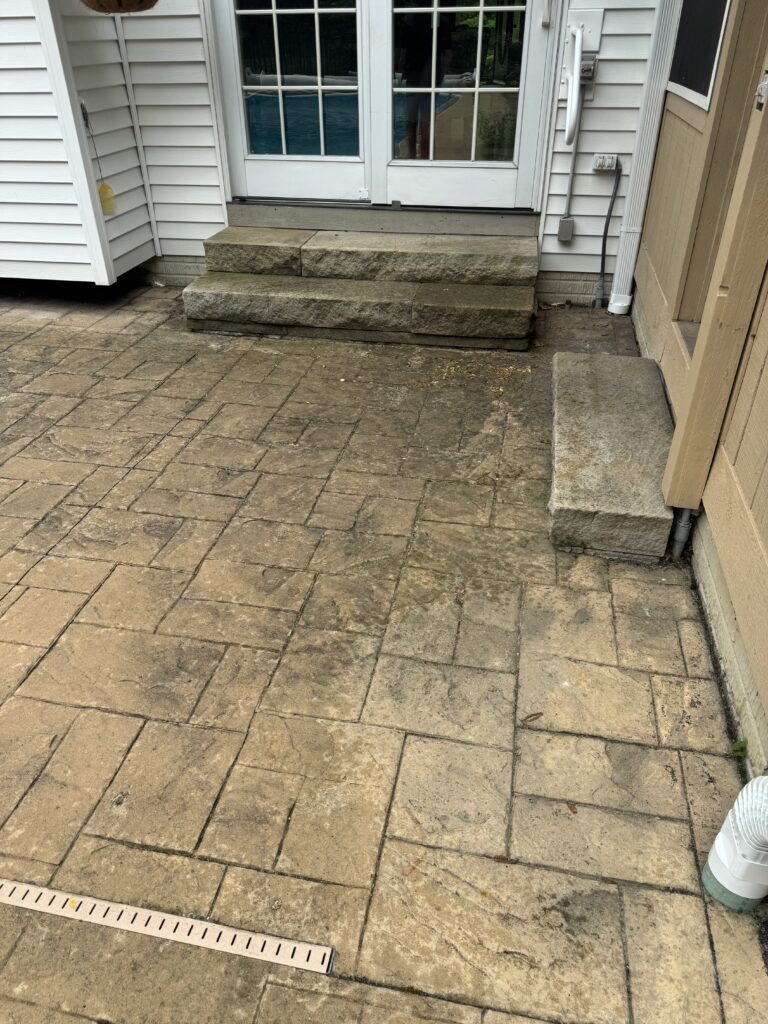 Backyard patio with three large stone steps up to white-framed glass doors; textured stone pavers extend outward. A measuring ruler lies along the edge of the patio. A hose and utility box are visible to the right.