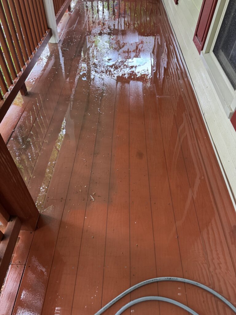Wet red wooden porch deck with reflections, a gray hose looping along the bottom edge, railing on the left and siding on the right after rain