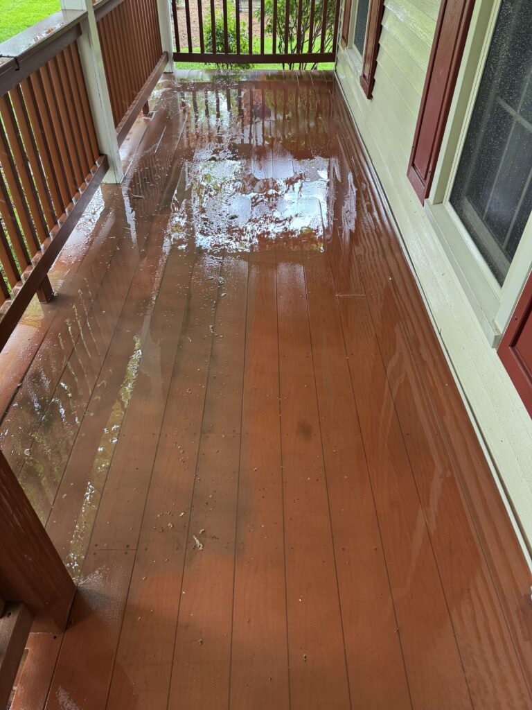 Wet brown wooden porch deck with standing water and reflections, railing on left and green lawn beyond. Useful for indicating a flooded outdoor porch scenario.