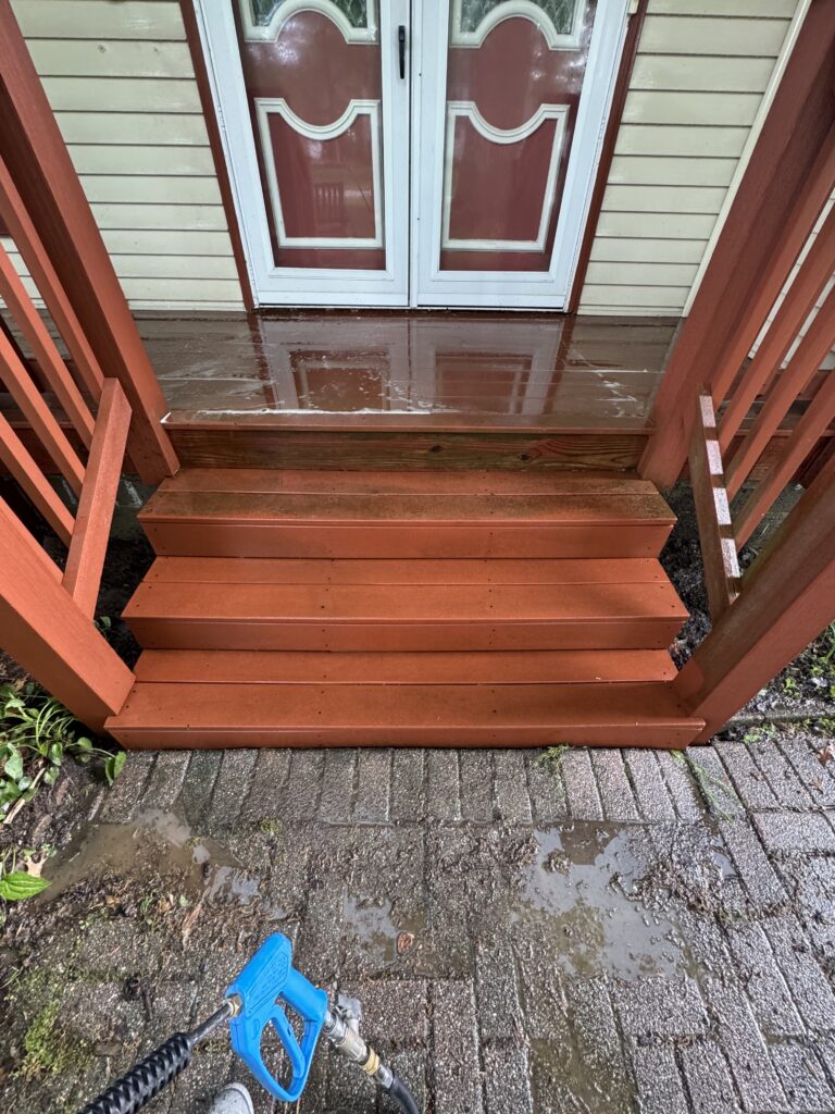 Wooden front steps stained orange-brown lead up to a white-framed double door with red panels; the wet steps have puddles and there’s a blue hose nozzle visible in the lower left foreground.