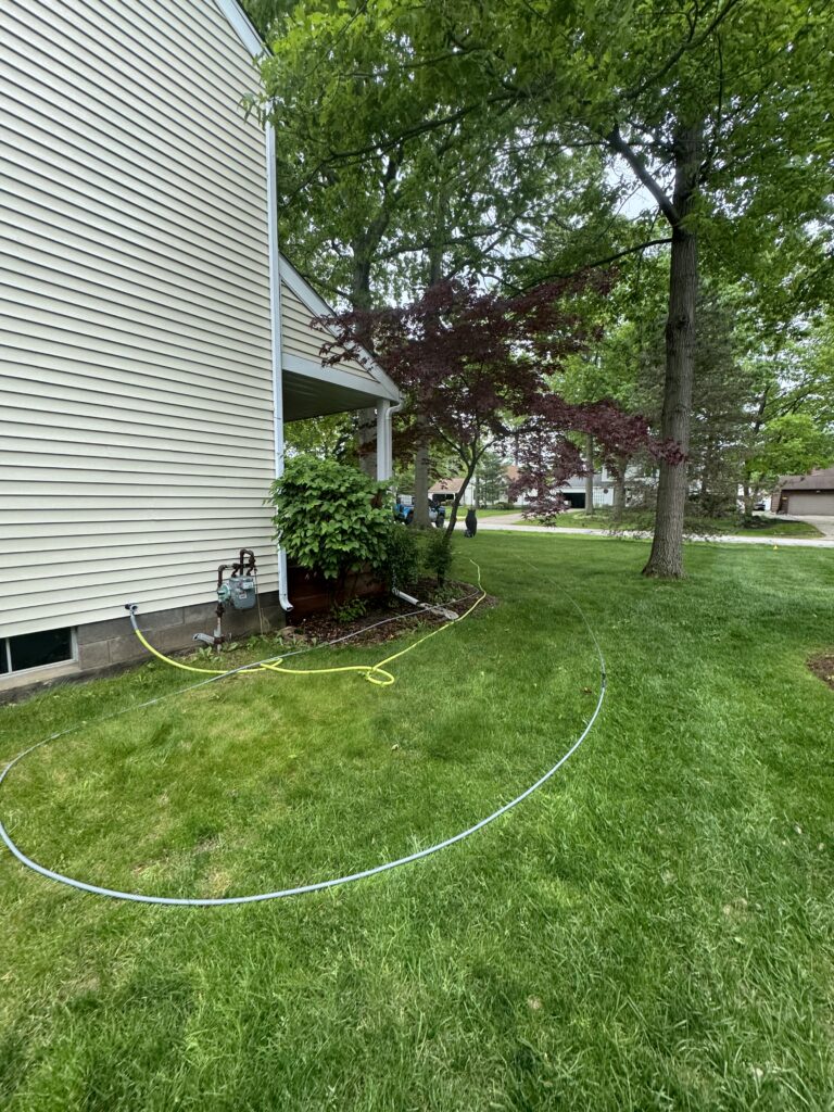 Backyard view of a beige house with a yellow hose looped on the green lawn near the foundation, trees in the background.