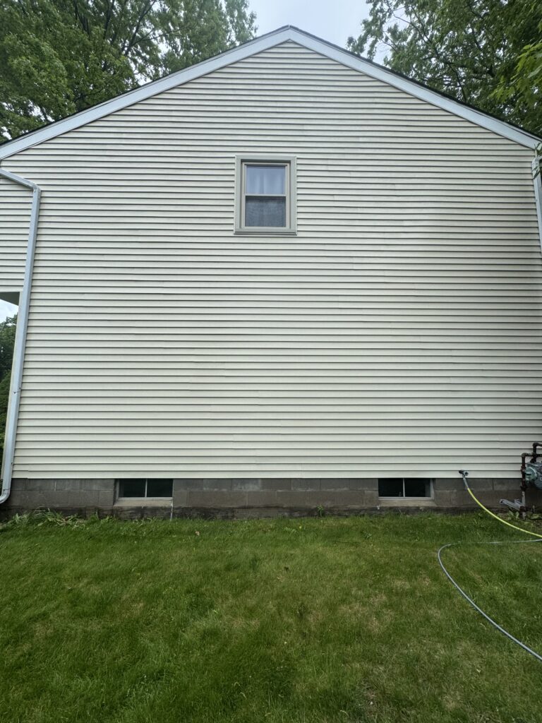 Side of a light-colored house with horizontal vinyl siding, a small rectangular basement window, and a grassy yard in front.