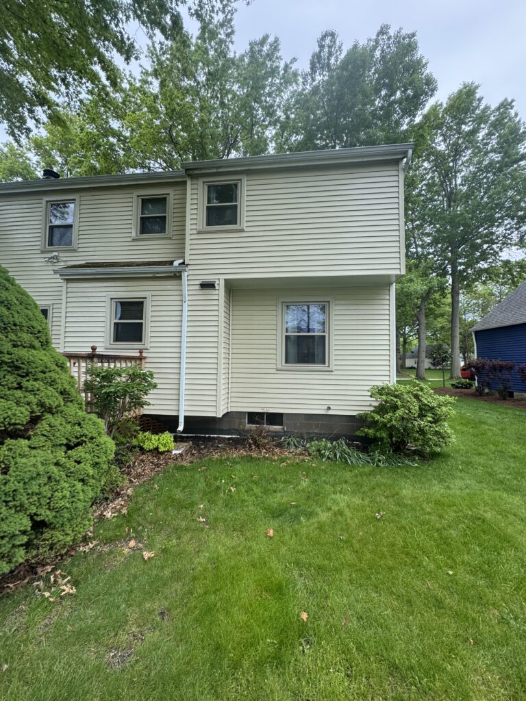 Two-story beige house with vinyl siding, several windows, white trim, and a well-kept green lawn with shrubs and trees in the background.