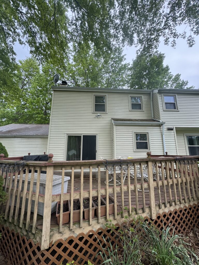 Backyard view of a beige two-story house with a sliding door, wooden deck, and lattice railing beneath a canopy of green trees.
