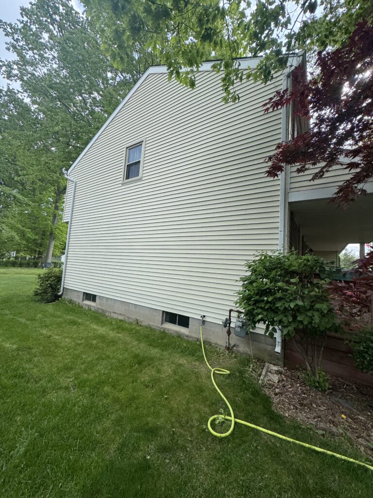 Side view of a beige vinyl-siding house with a small window, green lawn, and a yellow garden hose stretched across the grass.