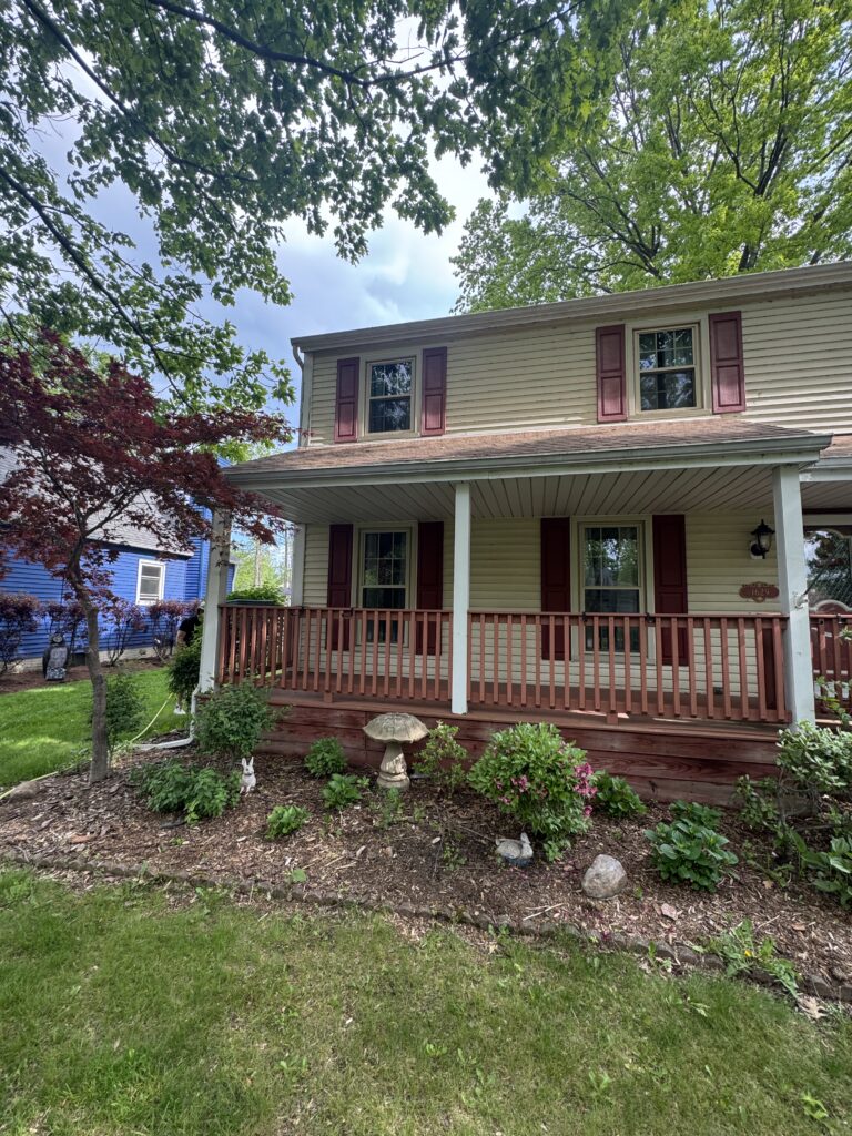 Two-story beige house with red shutters and a wooden front porch surrounded by green trees and a small garden bed in front.