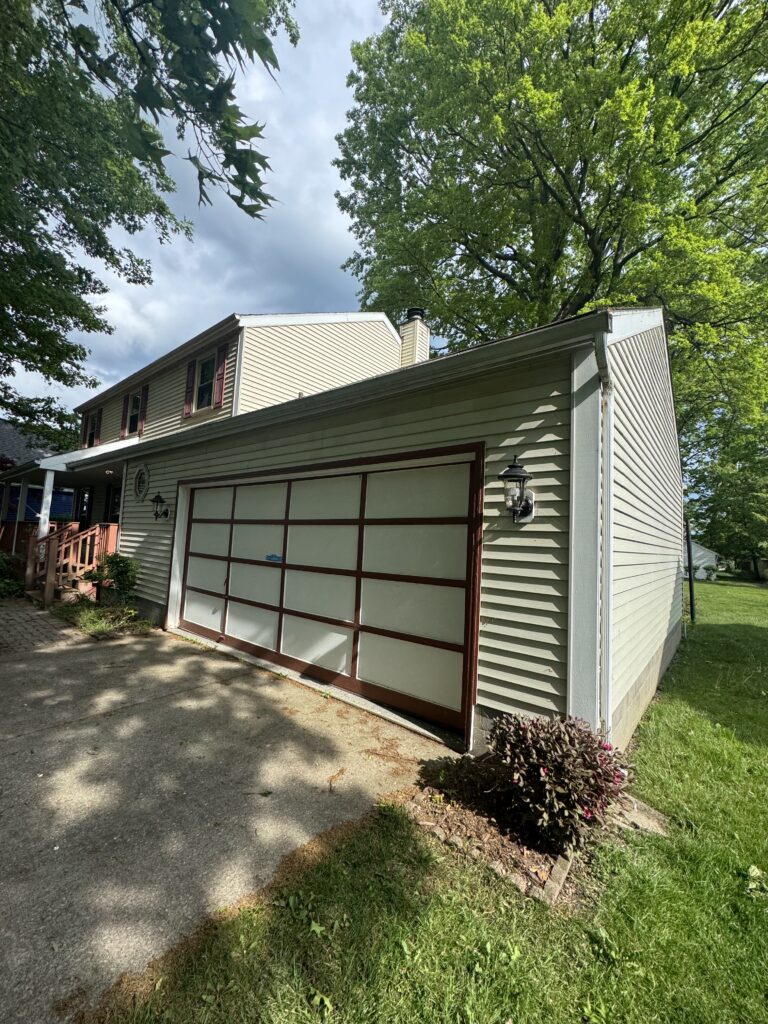 Front view of a suburban house with a large beige garage door, surrounded by green trees and a paved driveway.
