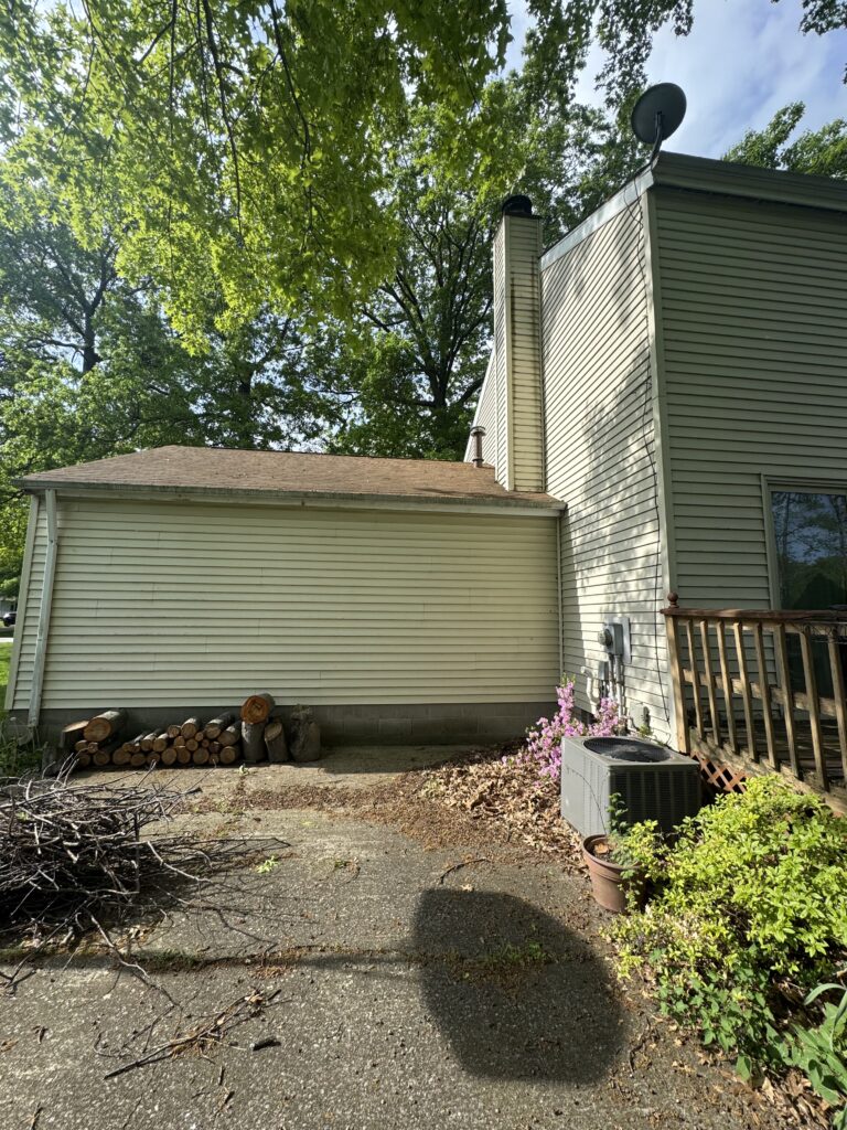 Backyard scene with beige house siding, stacked firewood, and a tangle of branches on the ground near an outdoor air conditioner unit and pink flowers.