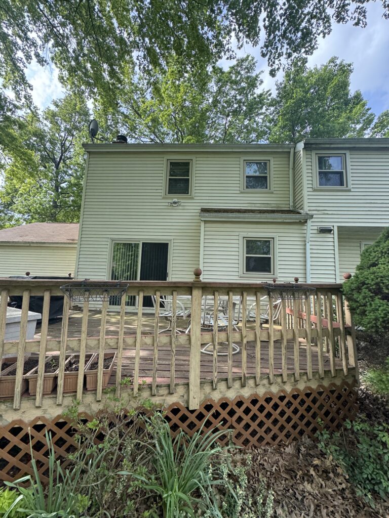 Backyard with a beige two-story house, wooden deck and railing, and leafy trees overhead.