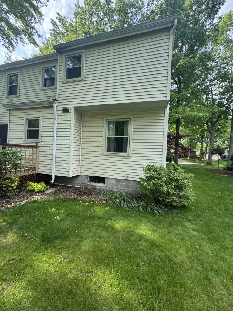 Two-story beige house with horizontal siding, several windows, and a small deck area, surrounded by a green lawn and trees across the yard.