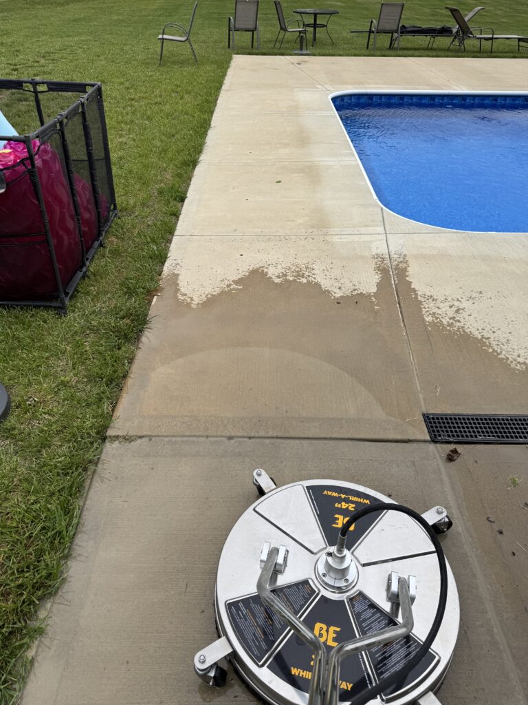 Poolside scene with a large circular pool reel on the concrete deck by a blue pool, hose attached, and lawn chairs in the background on the grass.