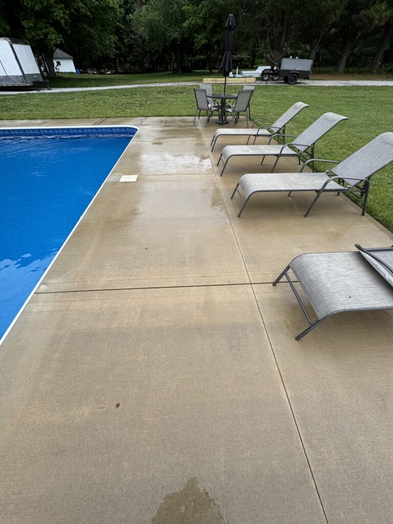 Outdoor poolside scene with blue pool, beige concrete deck, and a row of gray lounge chairs nearby.