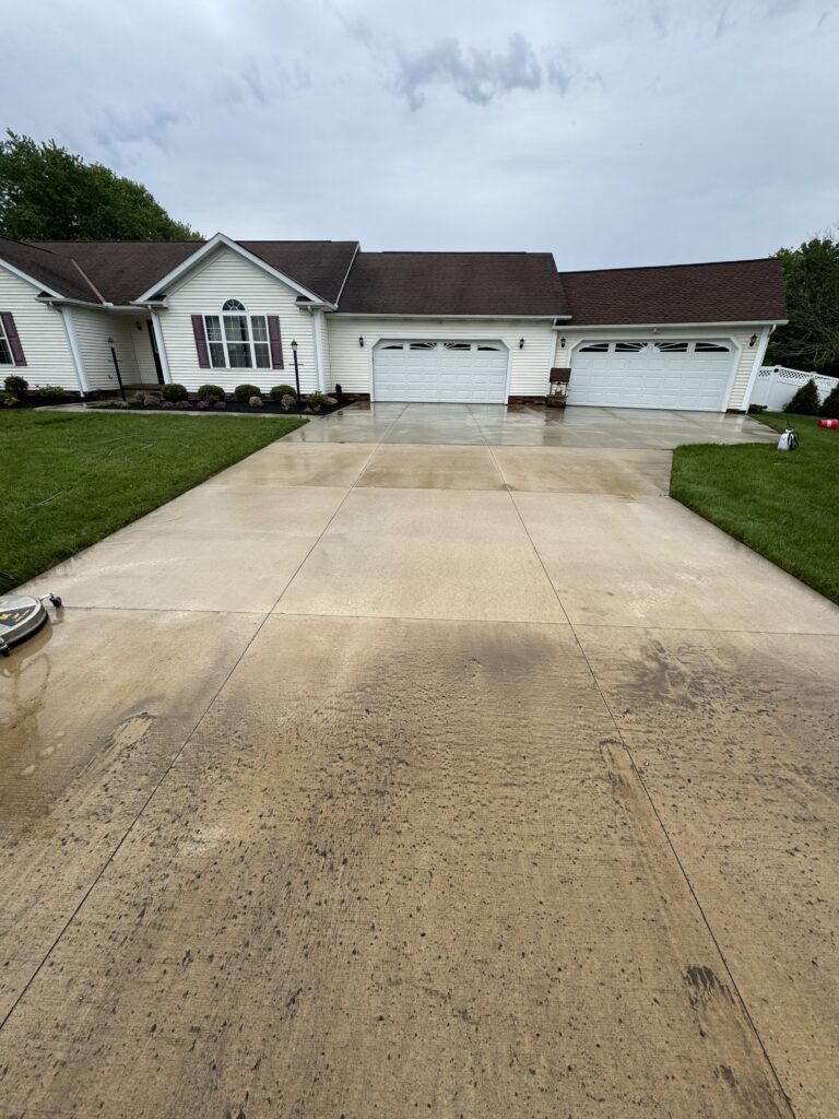 Large white suburban house with a three-car garage and wet concrete driveway after rain, cloudy sky above, green lawn on both sides.