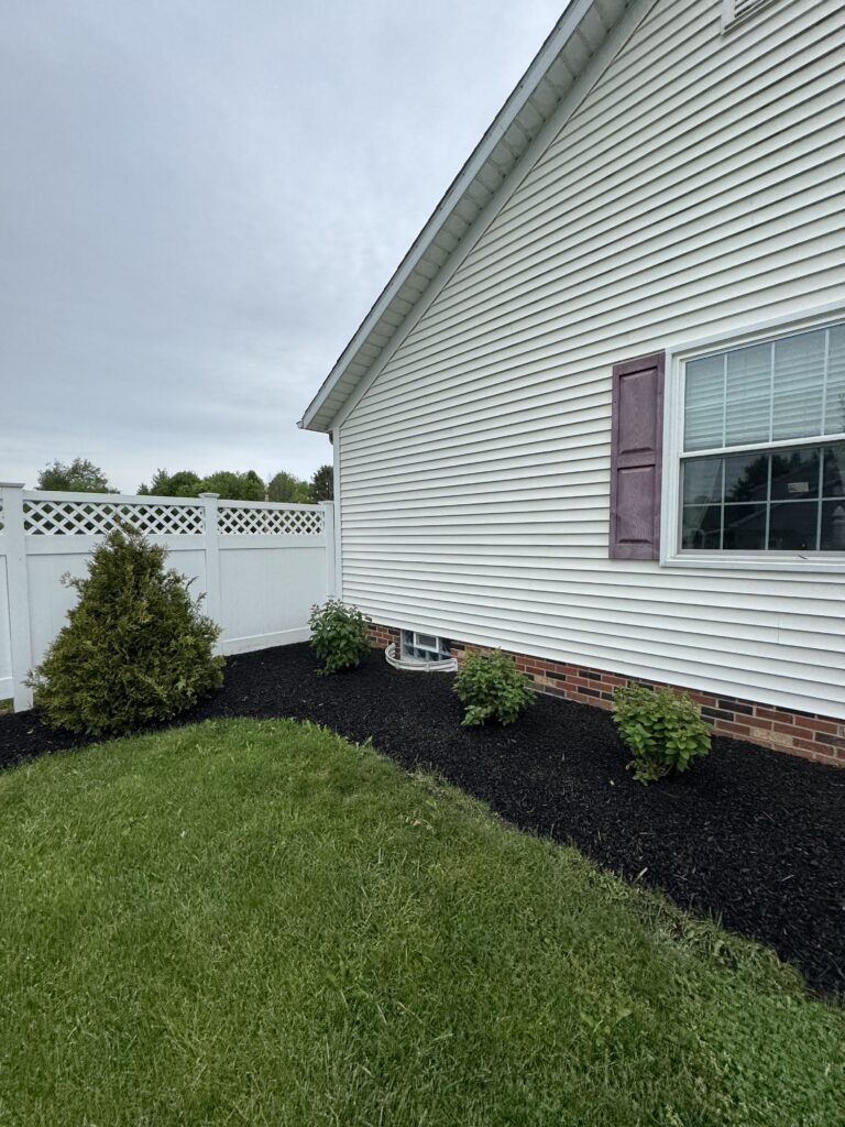 Side view of a white house with vinyl siding, a window with burgundy shutters, and a mulched garden bed along the foundation.