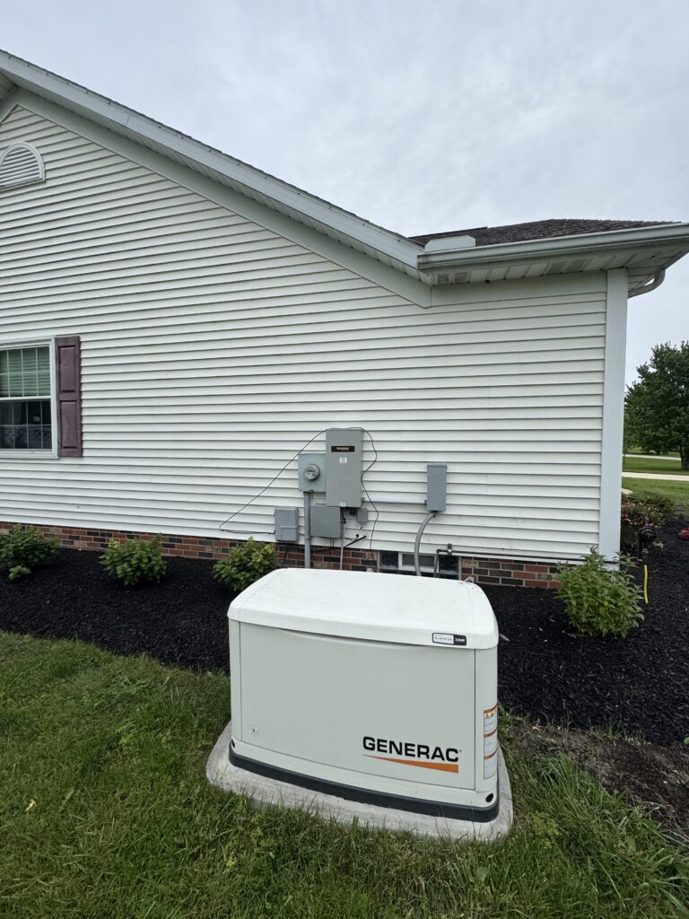 Exterior view of a Generac backup generator mounted on a concrete pad beside a white siding house with a brick foundation and landscaped mulch bed, ready to provide power during outages