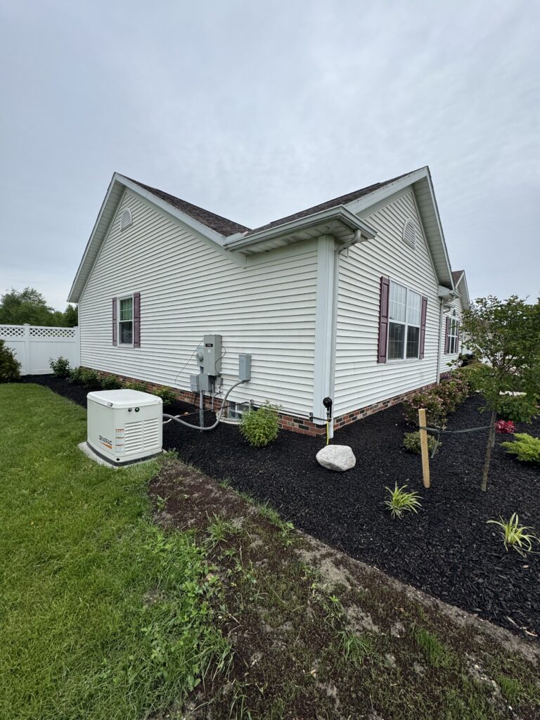 Side view of a white vinyl-sided house with maroon shutters, an outdoor air conditioner unit, and a mulched yard with small plants.