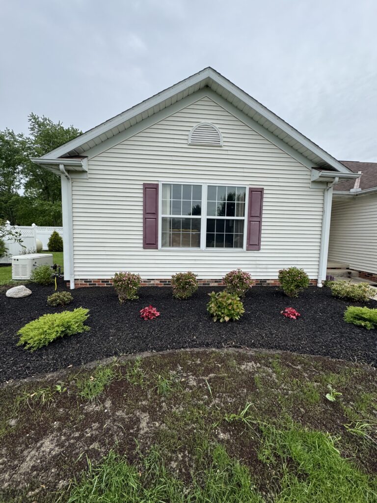 Front view of a beige house with a gabled roof, white trim, and pink shutters around a large window; dark mulched front yard with small shrubs.